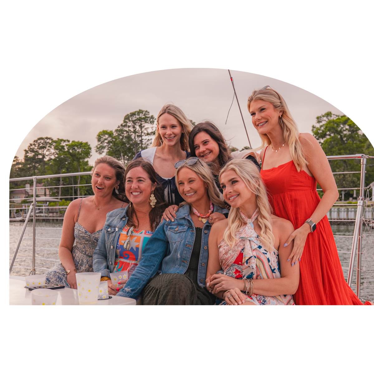 Seven women smiling on a boat with trees and water in the background.